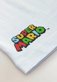 Biała bawełniana koszulka z multikolorowym graficznym nadrukiem "SUPER MARIO", z wyraźnym napisem i czarną obwódką na dolnej krawędzi.
