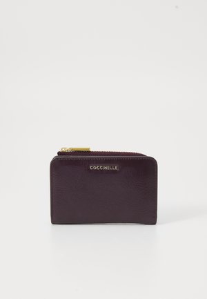 Portafoglio in pelle di un viola scuro con una texture sottile, dotato di una cerniera dorata e dettaglio del logo sul davanti. Forma rettangolare compatta.