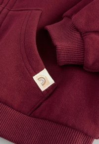 Sweatshirt bordeaux avec un tissu texturé, des poignets côtelés et une poche avant. Présente une étiquette tissée avec un logo circulaire.