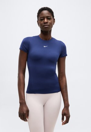 Kvinde iført en tætsiddende navyblå Nike kortærmet top og lyse beige leggings, stående mod en ensartet lys baggrund.