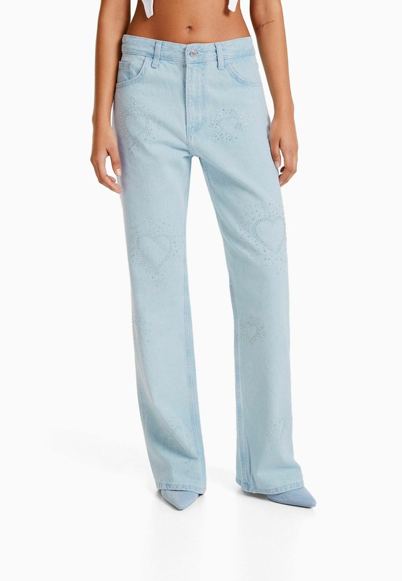 Jean en denim bleu clair de forme évasée, présentant des motifs de cœurs perlés et une ceinture standard avec une fermeture à bouton à l'avant.