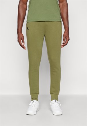 Joggers de color verde oliva hechos de una tela suave, con un corte ajustado, puños acanalados y un pequeño logo negro en el muslo izquierdo. Combinados con zapatillas blancas.