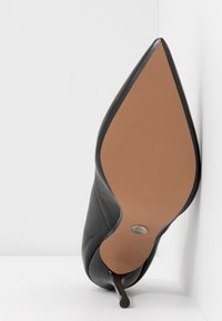 Chaussure à talon haut noire à bout pointu inclinée vers le haut, montrant une semelle lisse couleur beige et un talon aiguille fin sur un fond blanc.