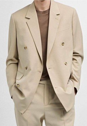 Beige zweireihige Anzugjacke mit Schildpattknöpfen, getragen über einem braunen Rundhalsshirt, Hände in den Taschen.