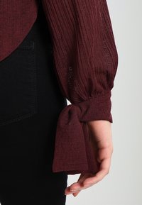 Marooni blusa ku tekstuuriga kangas, pikad varrukad, millel on kokku tõmmatud mansetid, seotud lipsuks, koos mustade pükstega.