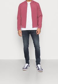 Muž v červené bomber bundě, bílé tričko s potiskem, tmavé úzké džíny a bílé tenisky s fialovými akcenty.