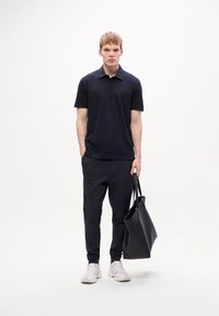 Joven de pie con camiseta polo negra, pantalones negros, zapatillas blancas, sosteniendo una bolsa tote negra contra un fondo blanco liso.