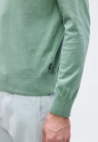 Homme portant un pull en maille vert clair à manches longues avec l'étiquette BOSS visible, associé à un jean bleu clair, main posée le long du corps.
