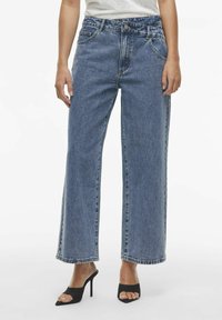Vaqueros de mezclilla azul de cintura alta y perneras anchas con un lavado claro, que cuentan con cinco bolsillos y detalles de costura en el dobladillo. Combinados con sandalias negras de tacón.