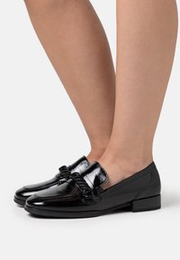 Mocassini in pelle verniciata nera con accentuazione a fiocco testurizzato, tacco basso a blocco e superficie liscia, che mostrano un design classico e formale.