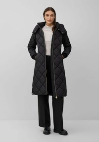 Femme portant un long manteau matelassé noir avec capuche, sur un col roulé blanc et un pantalon noir à jambes larges, se tenant devant un fond gris uni.