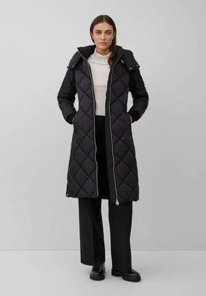 Woman wearing a long black quilted coat with hood over a white turtleneck and black wide-leg pants, standing against a plain gray background.