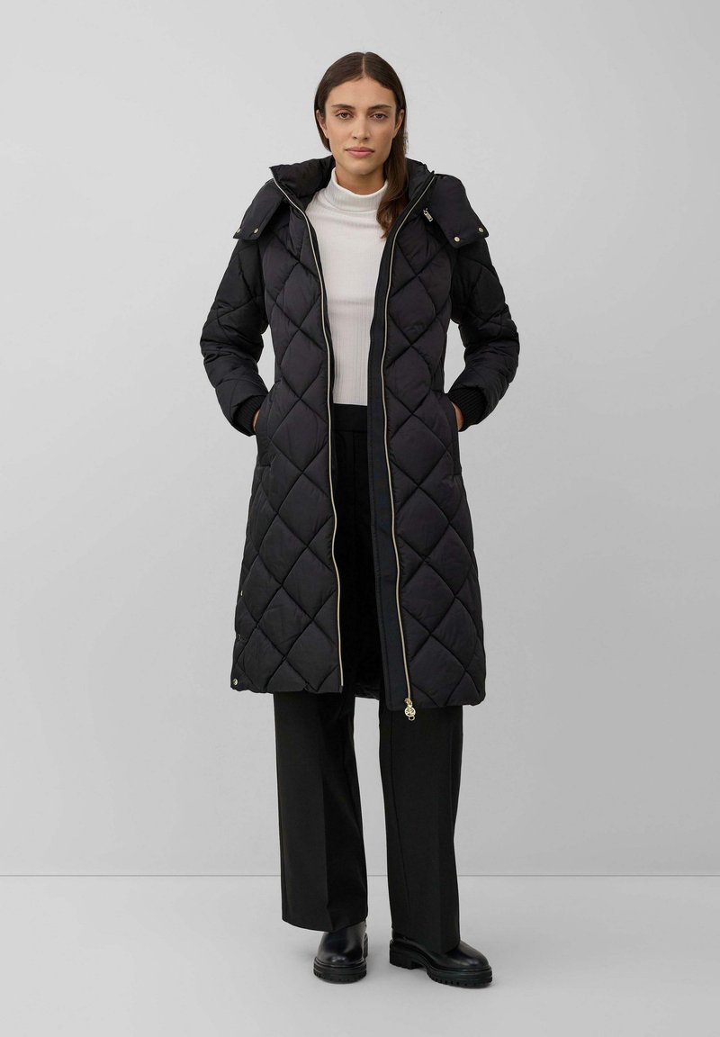 Femme portant un long manteau matelassé noir avec capuche, sur un col roulé blanc et un pantalon noir à jambes larges, se tenant devant un fond gris uni.
