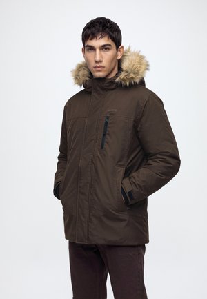 Homme portant un manteau d’hiver marron avec une capuche bordée de fourrure, les mains dans les poches, debout devant un fond clair uni.