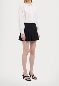 Valkoinen nappipaita, jossa on puffattuja hihoja, yhdistetty mustaan laskostettuun hameeseen ja mustiin remmissandaaleihin. Yksinkertainen ja rakenteellinen muotoilu.