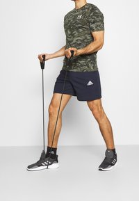 Kamuflážová krátká rukávová košile se zelenými tóny, námořnické šortky s bílým logem Adidas, šedé sportovní boty a fitness odporové gumy.