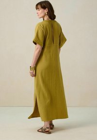 Robe maxi jaune moutarde avec des manches courtes, tissu texturé, ouverture dans le dos et fentes latérales. Complétée par un bracelet et des sandales imprimé animal.