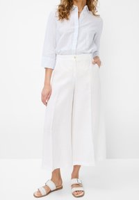 Pantaloni larghi bianchi in lino con chiusura frontale a bottone e tasche, abbinati a una camicia a righe azzurre con maniche lunghe e sandali piatti.