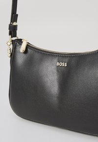 Borsa a tracolla in pelle nera con forma curvata. Presenta una cerniera dorata e dettagli in oro, logo "BOSS" in rilievo sulla parte frontale e tracolla regolabile.