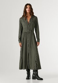 Μακρυμάνικο φόρεμα με κουμπιά σε πράσινο και μαύρο καρώ σχέδιο, τσάκιση στη μέση, με φαρδιά φούστα. Φοριέται με μαύρες γαλότσες μέχρι τον αστράγαλο.