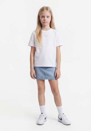 Junges Mädchen mit langen blonden Haaren, das ein weißes T-Shirt, einen blauen Jeansrock, weiße Nike-Socken und weiße Nike-Sneaker trägt und vor einem weißen Hintergrund steht.