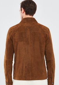 Bruine suède jas met een kraag, achternaden en lange mouwen. Zachte textuur met knopenaccenten bij de manchetten.