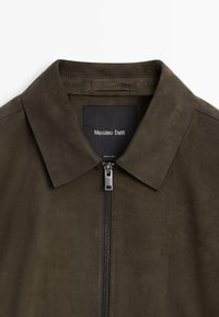 Brun mockakina med platt krage, framsida med dragkedja och en svart etikett med texten "Massimo Dutti" inuti. Mjuk textur och minimalistisk design.