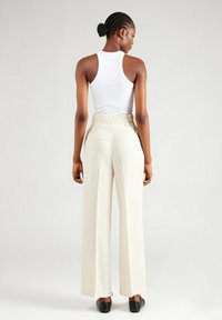 Hoge taille crèmekleurige broeken met een wijde pijp, voorzien van riemlussen en zijzakken. G gedragen met een witte tanktop en zwarte schoenen.