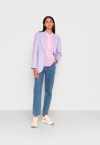 Blazer oversized lilás claro sobre uma camisa rosa, combinado com jeans de perna reta azuis e ténis rosa claro com detalhes brancos.