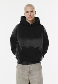 Felpa nera con un design a gradiente, realizzata in materiale morbido, con tasca a marsupio e cappuccio con cordini. Indossata con jeans grigio chiaro.