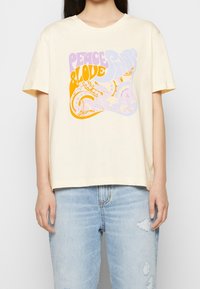 Mujer vistiendo una camiseta crema con texto en púrpura y naranja de "Paz y Amor" y diseño de paisley, combinada con jeans desgastados de color azul claro.