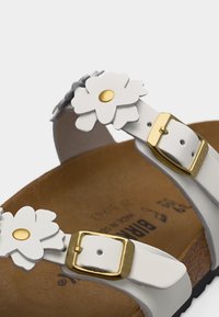 Λευκό σανδάλι με δύο χρυσά αγκράφες και λευκές διακοσμήσεις λουλουδιών στα λουριά, διαθέτει καφέ πάτο.