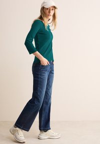 Tyrkysový dlouhý rukáv, světle modré široké džíny a bílé tenisky. Model má na sobě bílou čepici. Čisté, minimalistické pozadí.