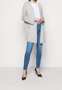 Cárdigan gris de punto con mangas anchas y dos bolsillos frontales, llevado sobre una camiseta blanca y unos vaqueros ajustados azules, acompañado de tacones negros.