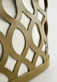 Tissu beige avec une superposition en cuir vert olive en forme de treillis, présentant des découpes ovales et en losange, cousu à la base sur un fond clair.