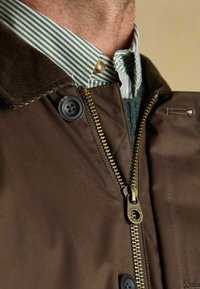 Brązowa kurtka zewnętrzna z kołnierzem sztruksowym, wyposażona w mosiężny zamek błyskawiczny, czarny guzik oraz zielono-biały pasek pod kołnierzem.