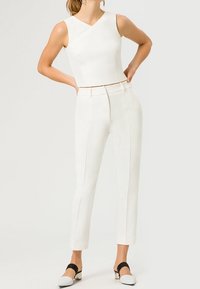 Donna che indossa un top corto bianco senza maniche, pantaloni bianchi a vita alta e scarpe bianche con tacco basso e cinturini neri, in piedi con le mani sui fianchi.
