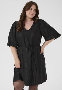 Musta mekko, jossa on teksturoitu kiilto, V-pääntie, lyhyet puffihihat ja kankainen nauha vyötäröllä, päättyen juuri polven ylle.