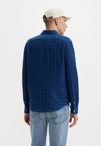 Camicia button-up blu navy con un motivo testurizzato e sottili quadrati azzurri. Maniche lunghe con polsini abbottonati. Abbinata a jeans in denim chiaro.