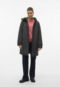Donkergrijze parka met capuchon, een rits aan de voorkant en drukknoopjes, gedragen over een roze sweater en donkere jeans. Zwarte schoenen met een gestructureerde afwerking.