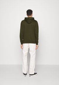 Hombre vestido con una sudadera verde oscuro, pantalones blancos y zapatillas negras, de pie frente a una pared blanca con las manos relajadas a los lados.