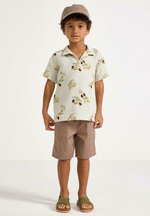 Poika, jolla on beige kukkakuvioinen lyhythihainen paita, ruskeat shortsit, vaaleanruskea lippalakki ja vihreät sandaalit, seisoo valkoista taustaa vasten.