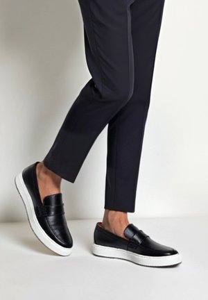 Black slip-on leather sneakers with white soles worn with black cropped trousers, showing ankles and partial legs in a walking pose.