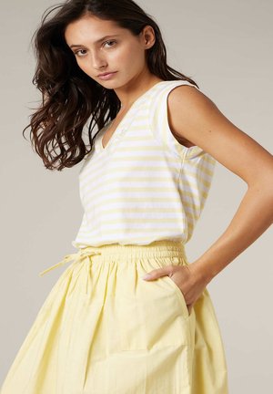 Femme aux cheveux bruns portant un haut sans manches rayé jaune et blanc et une jupe jaune taille haute avec une poche, posant avec une main dans la poche.