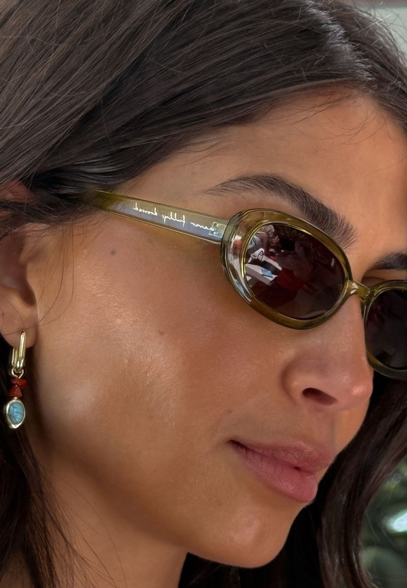 Žalios spalvos ovalūs saulės akiniai su tamsiais lęšiais, plonais metaliniais antakiais, ant kurių išgraviruotas logotipas, derinami su auksiniu ir raudonu kabančiu auskaru.