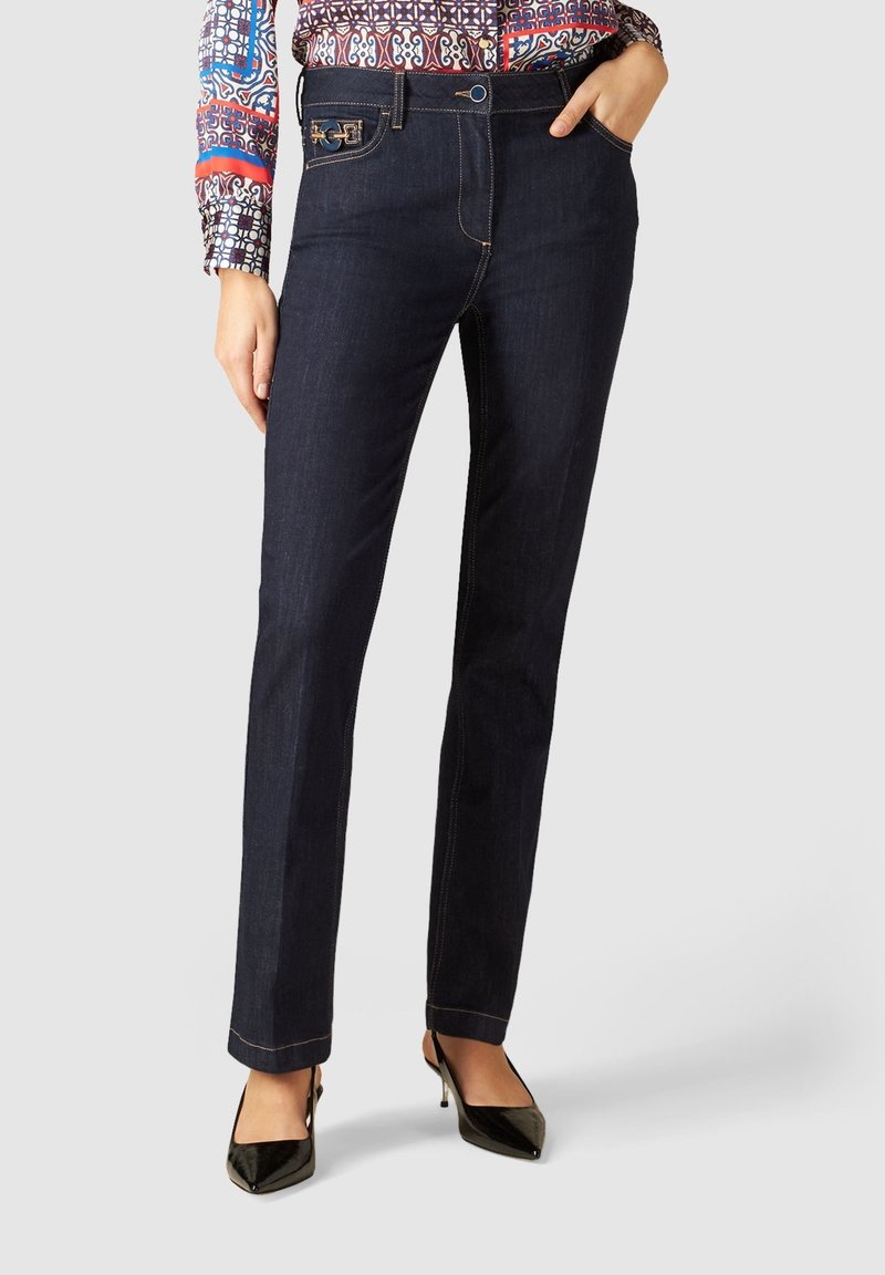 Femme portant un jean droit bleu foncé avec une chemise à manches longues à motifs, des escarpins noirs à bout pointu, les mains dans les poches.