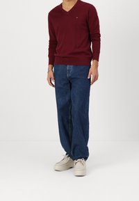 Pull col en V bordeaux, jean bleu décontracté à coupe ample, et chaussures casual beige clair. Le pull présente des manches longues et un petit logo en accent.