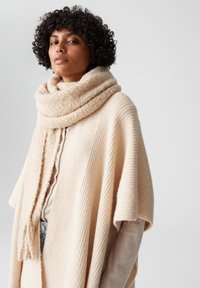 Poncho beige lavorato a maglia con texture a coste, dotato di una sciarpa morbida e pelosa. Il design include una vestibilità ampia e un'apertura frontale.
