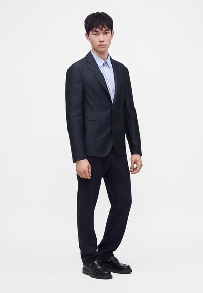 Homme portant une veste de costume noire ajustée, une chemise bleu clair, un pantalon noir et des chaussures en cuir noir, debout devant un fond blanc uni.