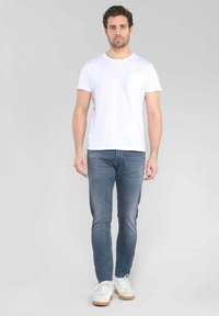 Una camiseta blanca de algodón con un bolsillo en el pecho, combinada con unos jeans ajustados azules y zapatillas blancas con suelas beige, de pie frente a un fondo gris.